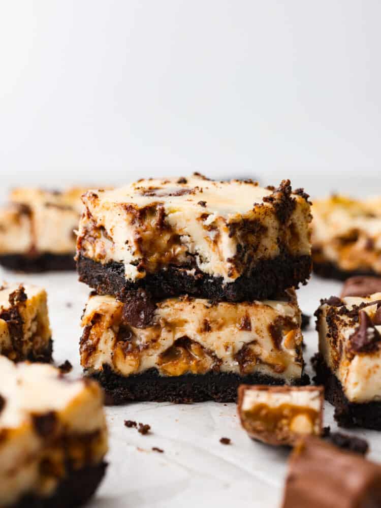 With a rich oreo crust and Snickers-filled cheesecake topping, these Snickers cheesecake bars are one of the best desserts out there! They're creamy, chocolatey, and sure to be loved by everyone! This is your sign to make a double batch. Closeup of 2 Snickers cheesecake bars stacked on top of each other.