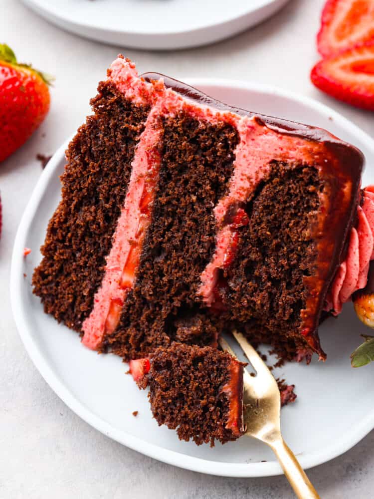 Impress your guests with this strawberry chocolate cake that is both gorgeous and decadently delicious. Rich layers of dark chocolate cake covered in sweet homemade strawberry frosting and fresh sliced strawberries...a match made in heaven! A close up photo of a slice of strawberry chocolate cake on a gray plate. A fork is lifting up a bite of cake. Sliced strawberries are garnished on the side of the plate.