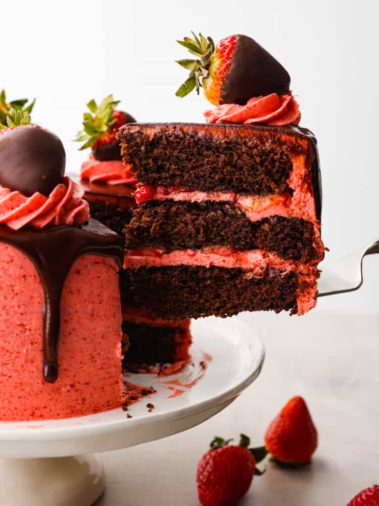 Impress your guests with this strawberry chocolate cake that is both gorgeous and decadently delicious. Rich layers of dark chocolate cake covered in sweet homemade strawberry frosting and fresh sliced strawberries...a match made in heaven! Side photo of a slice of strawberry chocolate cake being lifted off the cake platter.