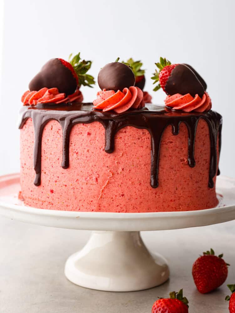 Impress your guests with this strawberry chocolate cake that is both gorgeous and decadently delicious. Rich layers of dark chocolate cake covered in sweet homemade strawberry frosting and fresh sliced strawberries...a match made in heaven! Front view of strawberry chocolate cake on a white cake stand and white background. Whole strawberries are garnished on the side of the cake stand.