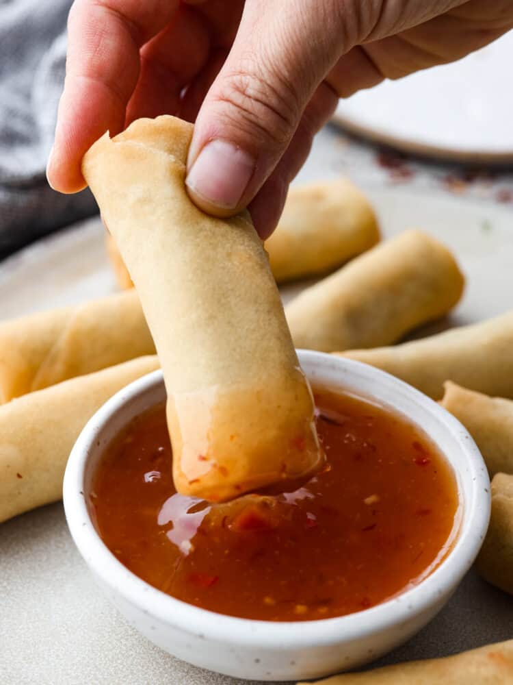 Homemade sweet chili sauce is the perfect dipping sauce to serve with chicken, seafood, and fried appetizers. A deliciously sweet and sour condiment full of Thai-inspired flavors, sugar, vinegar, and red chili paste. A side view of a small bowl full of sweet chili sauce with an egg roll being dipped into the sauce. The bowl of sauce is on a gray plate with egg rolls scattered all around the plate close to the sauce.