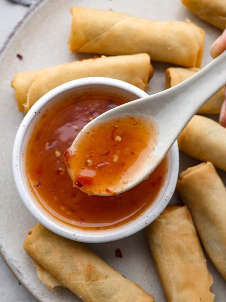 Homemade sweet chili sauce is the perfect dipping sauce to serve with chicken, seafood, and fried appetizers. A deliciously sweet and sour condiment full of Thai-inspired flavors, sugar, vinegar, and red chili paste. A top view of a small bowl full of sweet chili sauce and a spoon lifting out a spoonful of sauce. The bowl of sauce is on a gray plate with egg rolls scattered all around the plate close to the sauce.