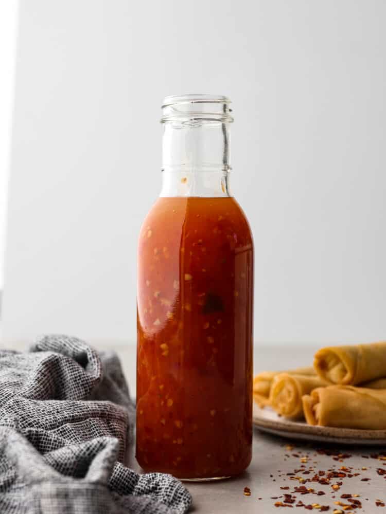 Homemade sweet chili sauce is the perfect dipping sauce to serve with chicken, seafood, and fried appetizers. A deliciously sweet and sour condiment full of Thai-inspired flavors, sugar, vinegar, and red chili paste. Side view photo of sweet chili sauce in a tall glass bottle with a gray kitchen towel and a plate of egg rolls next to the bottle. Red chili flakes are scattered on top of the gray countertop.