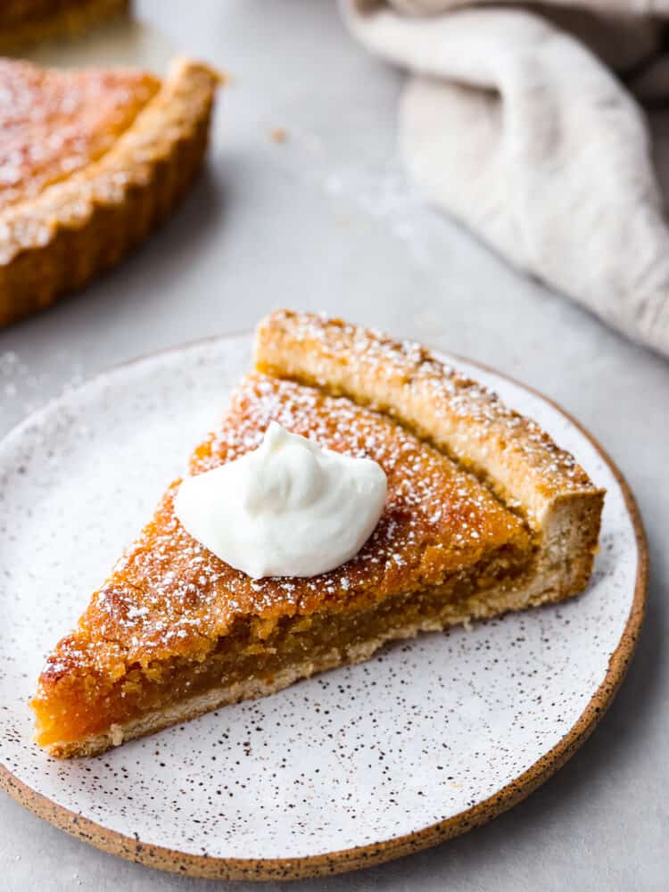 Treacle Tart is a buttery, creamy dessert brightened up by fresh lemon. The brown sugar shortbread crust is flaky and full of flavor. This tart is perfect for your holiday party or served with a cup of tea or coffee after dinner. Add a little whipped cream on top, and enjoy this delicious English dessert! A slice of treacle tart with a dollop of whipped cream on top on a stone-like plate.