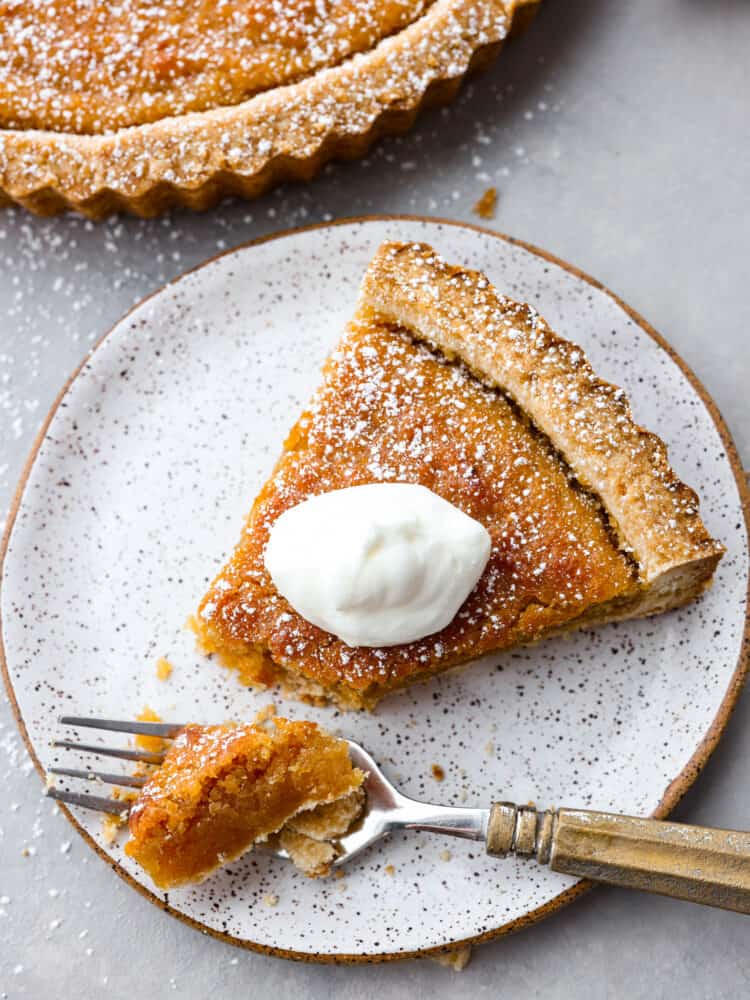 Treacle Tart is a buttery, creamy dessert brightened up by fresh lemon. The brown sugar shortbread crust is flaky and full of flavor. This tart is perfect for your holiday party or served with a cup of tea or coffee after dinner. Add a little whipped cream on top, and enjoy this delicious English dessert! A piece of tart with a scoop sitting on a spoon, topped with whipped cream and the full tart to the side.