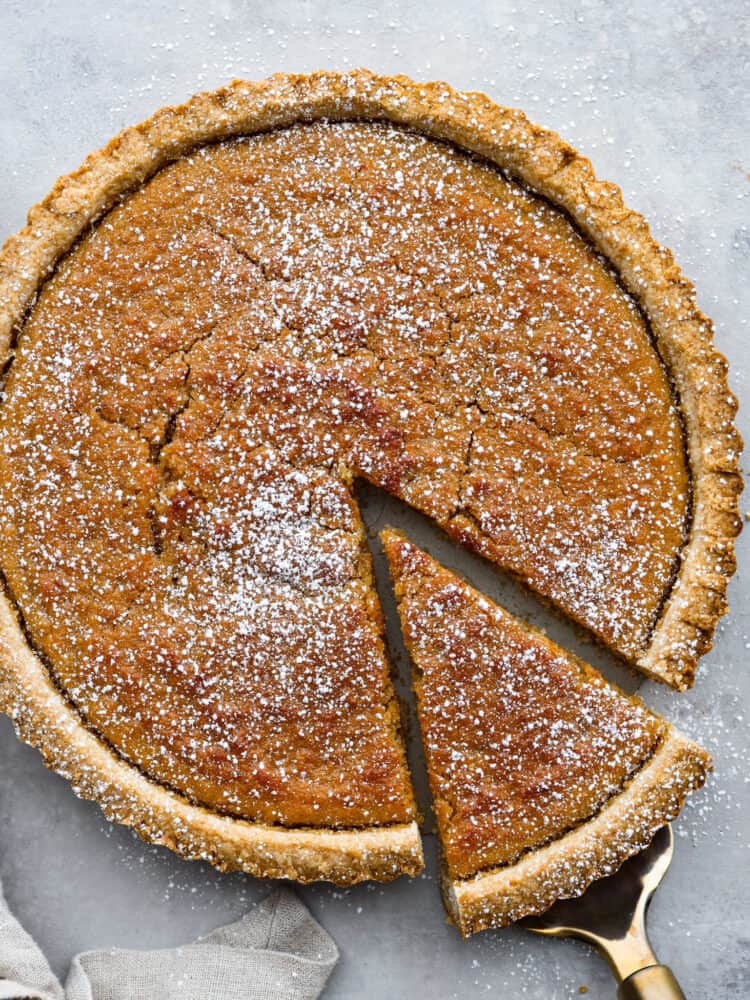 Treacle Tart is a buttery, creamy dessert brightened up by fresh lemon. The brown sugar shortbread crust is flaky and full of flavor. This tart is perfect for your holiday party or served with a cup of tea or coffee after dinner. Add a little whipped cream on top, and enjoy this delicious English dessert! Treacle tart with powdered sugar sprinkled on top with a slice being lifted out.