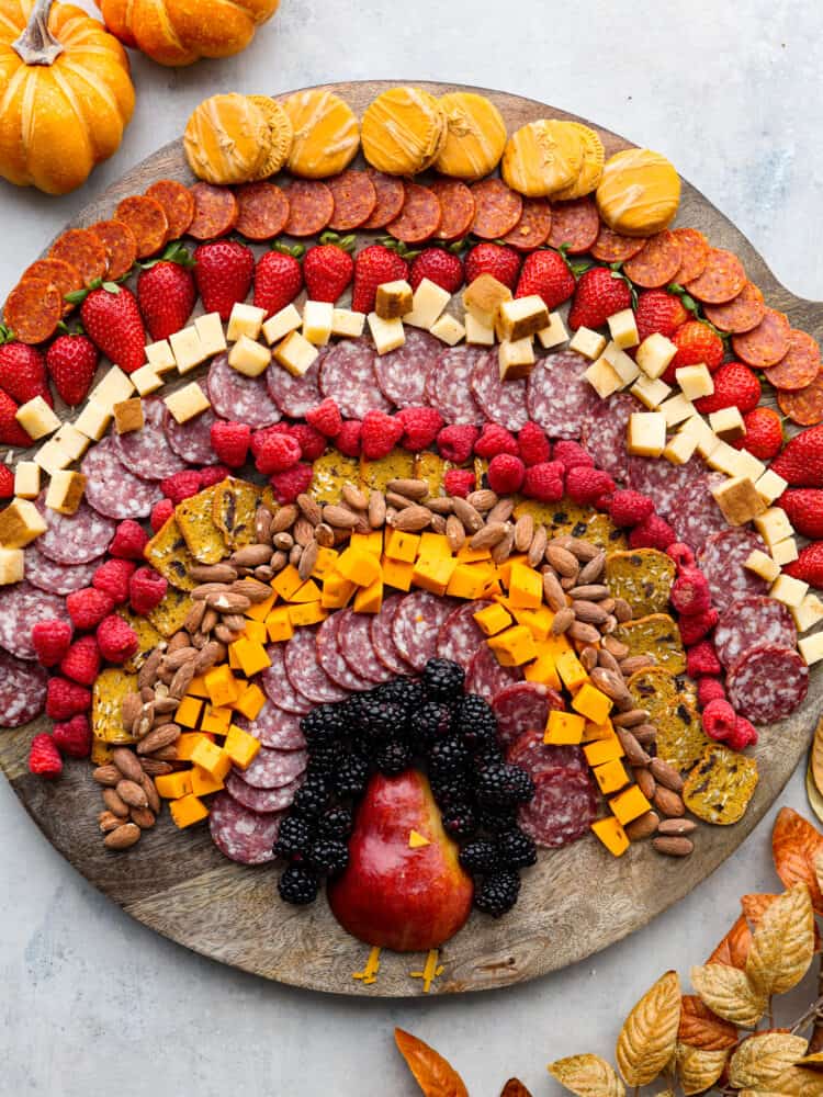 Gobble gobble your way to Thanksgiving dinner with the cutest turkey charcuterie board there ever was! Guests of all ages will love this fun way to enjoy a snack or appetizer full of meat, cheese, crackers, and lots of fruit! Overhead view of a turkey charcuterie board. Pumpkins and fall leaves are garnished around the wood board.