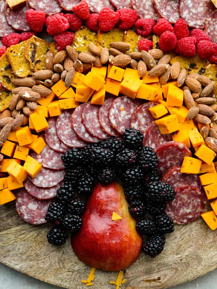 Gobble gobble your way to Thanksgiving dinner with the cutest turkey charcuterie board there ever was! Guests of all ages will love this fun way to enjoy a snack or appetizer full of meat, cheese, crackers, and lots of fruit! A close up view of the pear shaped turkey body. Fruit, salami, cheese, crackers, and nuts surround the turkey body.
