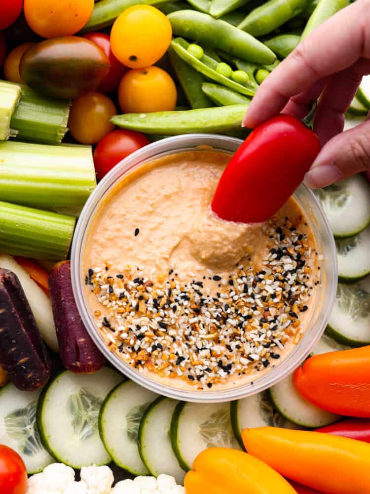 Every get-together needs a nutritious veggie charcuterie board full of colorful vegetables beautifully displayed all together. Perfect as an appetizer or side dish, this healthy board of fresh veggies is a hit served with a side of hummus and ranch dressing. Close up of a red bell pepper dipping into the hummus. Veggies surround the hummus.