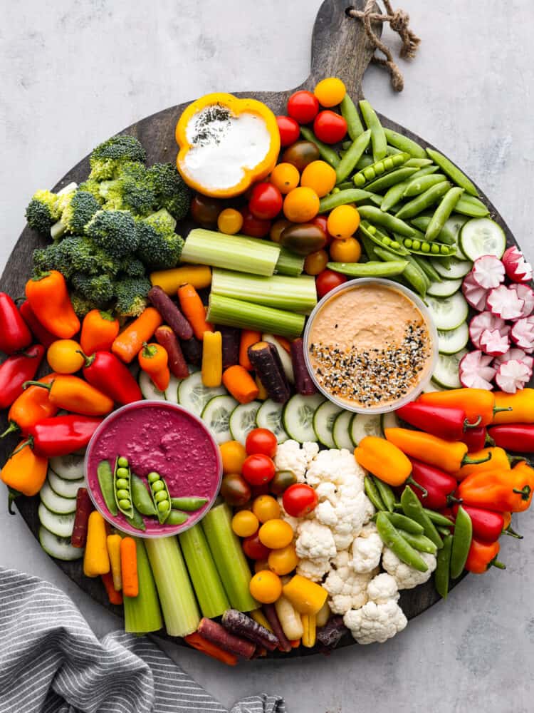 Every get-together needs a nutritious veggie charcuterie board full of colorful vegetables beautifully displayed all together. Perfect as an appetizer or side dish, this healthy board of fresh veggies is a hit served with a side of hummus and ranch dressing. Top view of a veggie charcuterie board on a large round wood board. A kitchen towel is styled next to it.