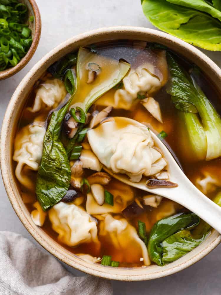 This homemade wonton soup is better than Chinese takeout and so easy to make! Enjoy the comforting flavors of tender wontons, vegetables, and warm broth seasoned with classic Chinese flavors of ginger, soy sauce, sesame oil, and garlic. Top view of wonton soup in a tan bowl with a white spoon lifting up a wonton dumpling. A tan towel and bowl of green onions are next to the bowl.