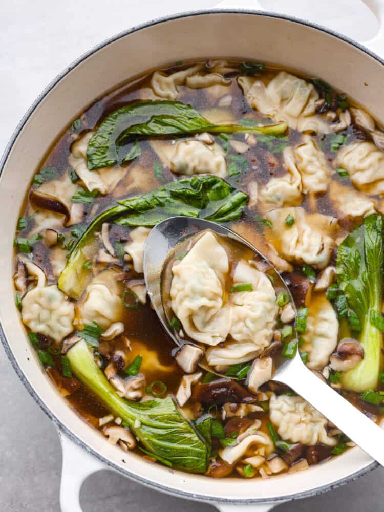 This homemade wonton soup is better than Chinese takeout and so easy to make! Enjoy the comforting flavors of tender wontons, vegetables, and warm broth seasoned with classic Chinese flavors of ginger, soy sauce, sesame oil, and garlic. Large white pot full of wonton soup. A large silver serving spoon is lifting the soup out of the pot.