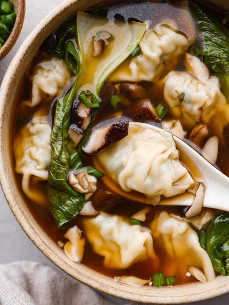 This homemade wonton soup is better than Chinese takeout and so easy to make! Enjoy the comforting flavors of tender wontons, vegetables, and warm broth seasoned with classic Chinese flavors of ginger, soy sauce, sesame oil, and garlic. Close up photo of wonton soup in a tan bowl with a white spoon lifting up a wonton dumpling. A tan towel and bowl of green onions are next to the bowl.