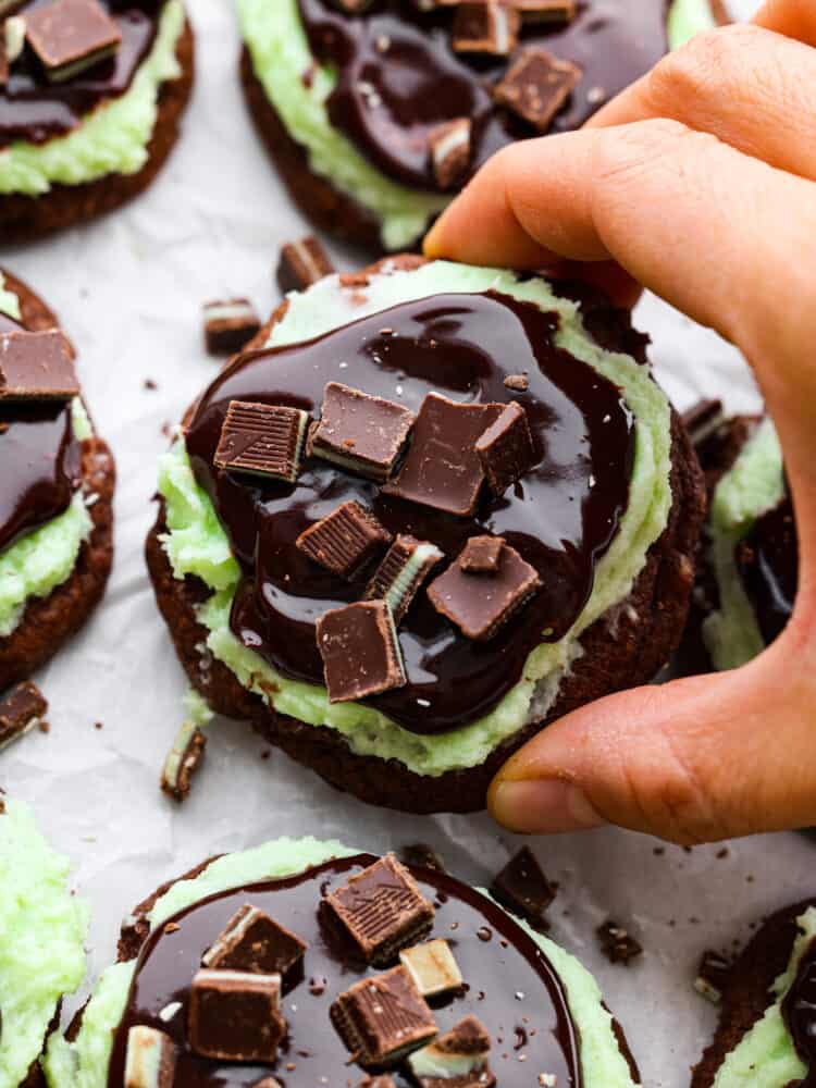 These Andes mint cookies are chewy and soft, topped with a lovely mint frosting and silky chocolate ganache. Andes mint candies sit on top and melt into the cookie as you chew, making it the perfect bite every time! A hand holding a chocolate cookie with other cookies in the background.