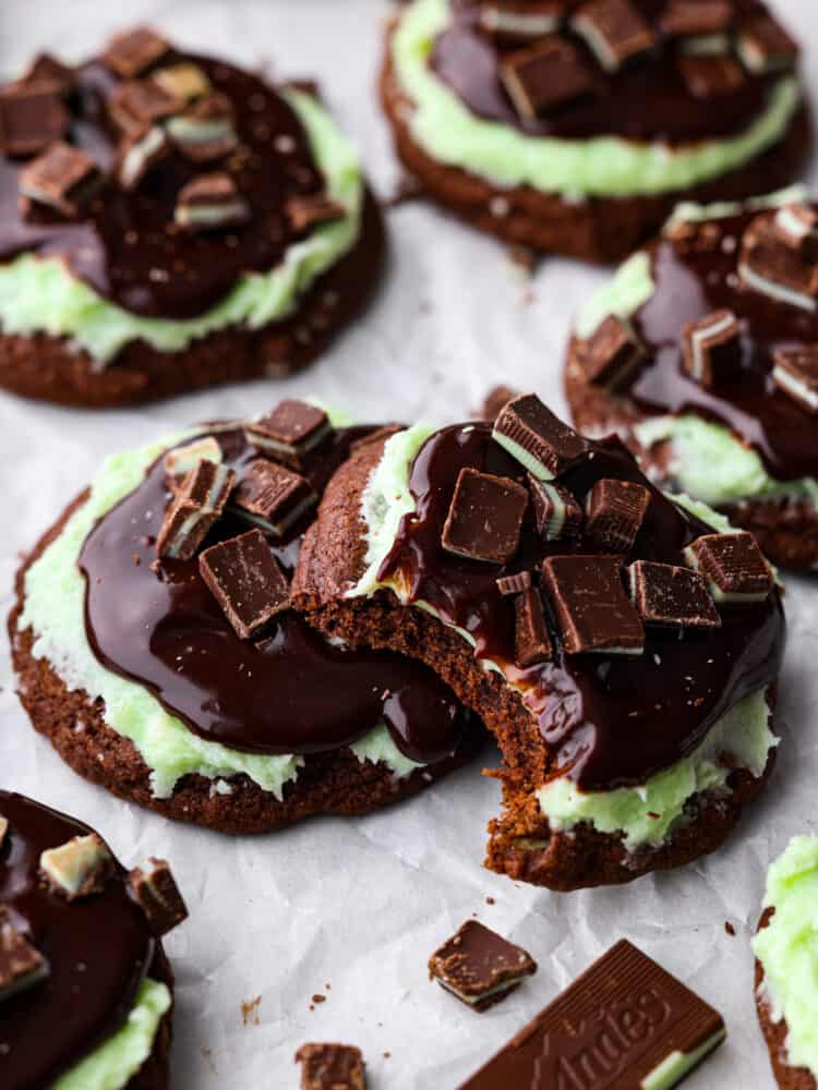 These Andes mint cookies are chewy and soft, topped with a lovely mint frosting and silky chocolate ganache. Andes mint candies sit on top and melt into the cookie as you chew, making it the perfect bite every time! Andes mint chocolate cookies on parchment paper, and one of the cookies has a bite taken out of it.