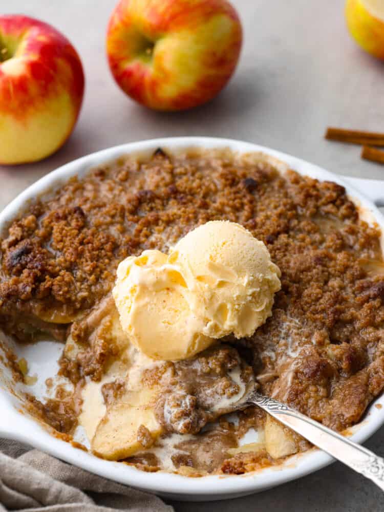 An Apple Brown Betty is the ultimate comfort dessert, filled with tender apples coated in a brown sugar-spiced crumble. With its warm, cozy flavors, this classic treat captures the essence of fall in every bite. Apple Brown Betty in a white dish with vanilla ice cream on top and scoops taken out. A metal spoon is sitting in the dish with apples in the background