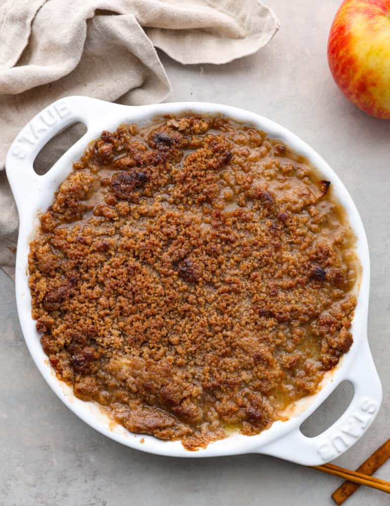 An Apple Brown Betty is the ultimate comfort dessert, filled with tender apples coated in a brown sugar-spiced crumble. With its warm, cozy flavors, this classic treat captures the essence of fall in every bite. A finished and whole Apple Brown Betty in a white baking dish with a tan tea towel next to it.