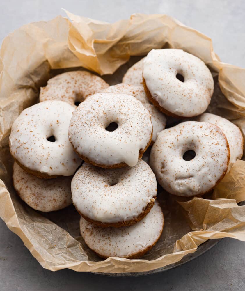 These baked apple cider donuts are rich, soft, and filled with spiced apple flavor! Topped with a creamy apple glaze, they almost look too good to eat! Theyโre the perfect addition to any fall get-together. Glazed apple cider donuts in a bowl lined with parchment paper.