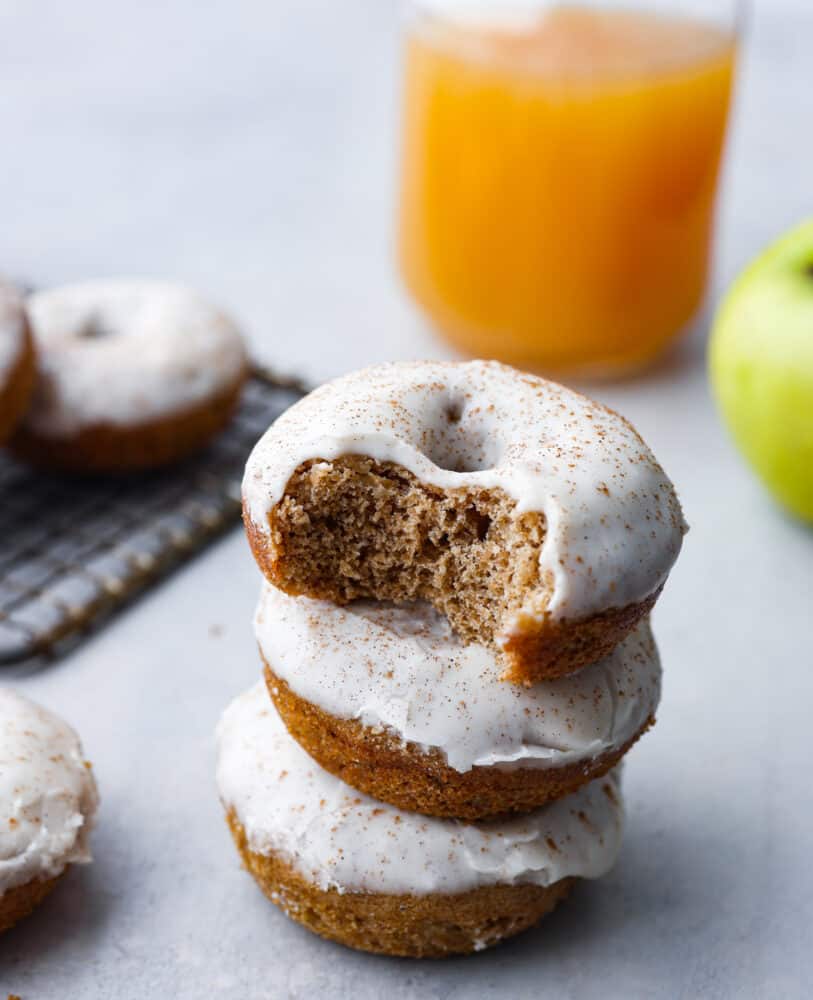 These baked apple cider donuts are rich, soft, and filled with spiced apple flavor! Topped with a creamy apple glaze, they almost look too good to eat! Theyโre the perfect addition to any fall get-together. A stack of 3 donuts. The top donut has a bite taken out of it.