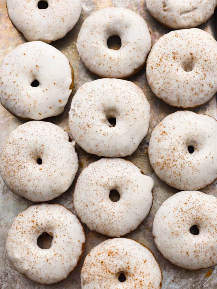 These baked apple cider donuts are rich, soft, and filled with spiced apple flavor! Topped with a creamy apple glaze, they almost look too good to eat! Theyโre the perfect addition to any fall get-together. Top-down view of baked apple cider donuts on parchment paper.