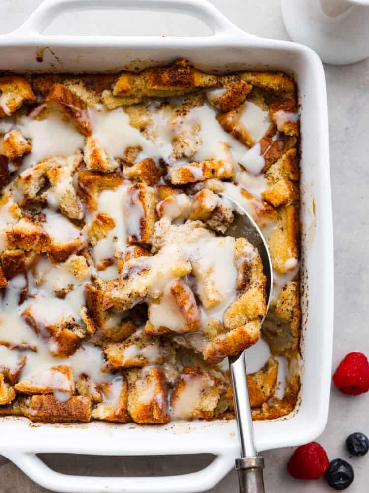 This Bread Pudding is a classic dessert that uses your leftover bread and a delicious vanilla sauce to make the best breakfast dessert you've ever had! It's frugal and cheap to make, but the flavor is out of this world divine. Bread pudding in a white pan with vanilla sauce on top and a metal spoon taking a scoop out.