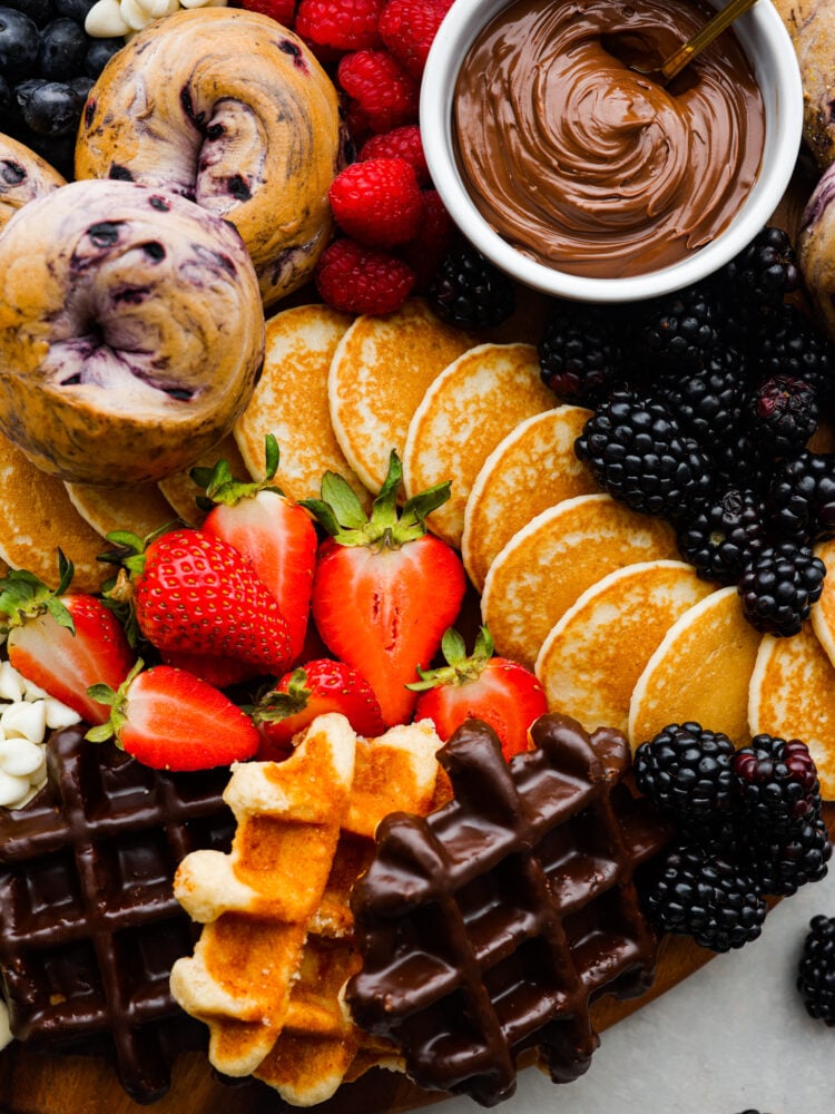 Wake up your family or guests to the world's CUTEST breakfast charcuterie board! Filled with everyone's favorite breakfast indulgences of waffles, pancakes, bagels, fruit, and all the decadent toppings your heart desires. Close up view of pancakes, waffles, and bagels on the breakfast charcuterie board. Colorful fruit and Nutella are scattered around the pancakes.