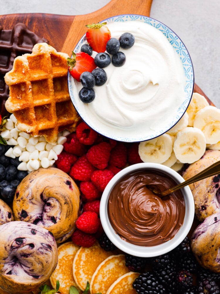 Wake up your family or guests to the world's CUTEST breakfast charcuterie board! Filled with everyone's favorite breakfast indulgences of waffles, pancakes, bagels, fruit, and all the decadent toppings your heart desires. Close up of whipped cream, Nutella, and fruit.