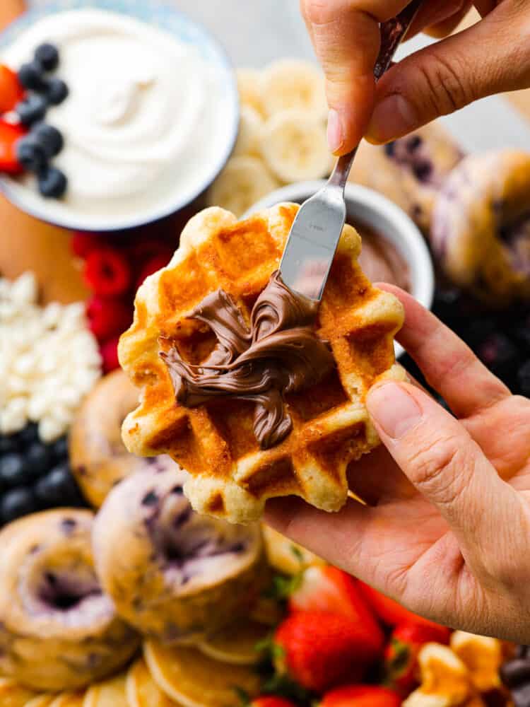 Wake up your family or guests to the world's CUTEST breakfast charcuterie board! Filled with everyone's favorite breakfast indulgences of waffles, pancakes, bagels, fruit, and all the decadent toppings your heart desires. Close up view of Nutella being spread on a waffle with the board of ingredients in the background.