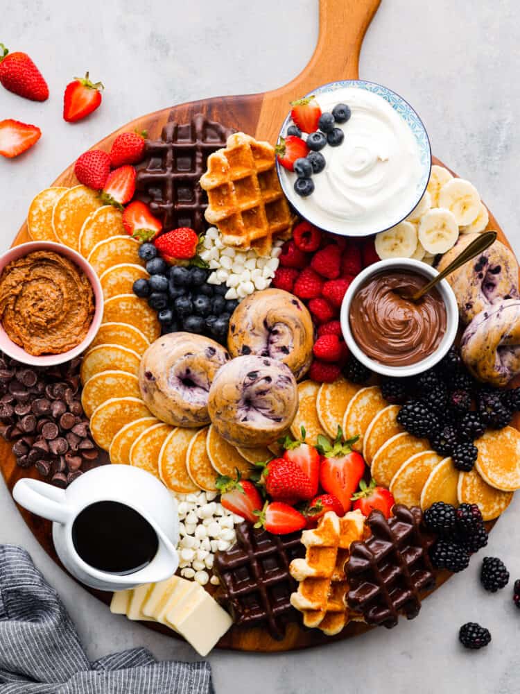 Wake up your family or guests to the world's CUTEST breakfast charcuterie board! Filled with everyone's favorite breakfast indulgences of waffles, pancakes, bagels, fruit, and all the decadent toppings your heart desires. Overhead view of the breakfast charcuterie board with fruit scattered next to the board.