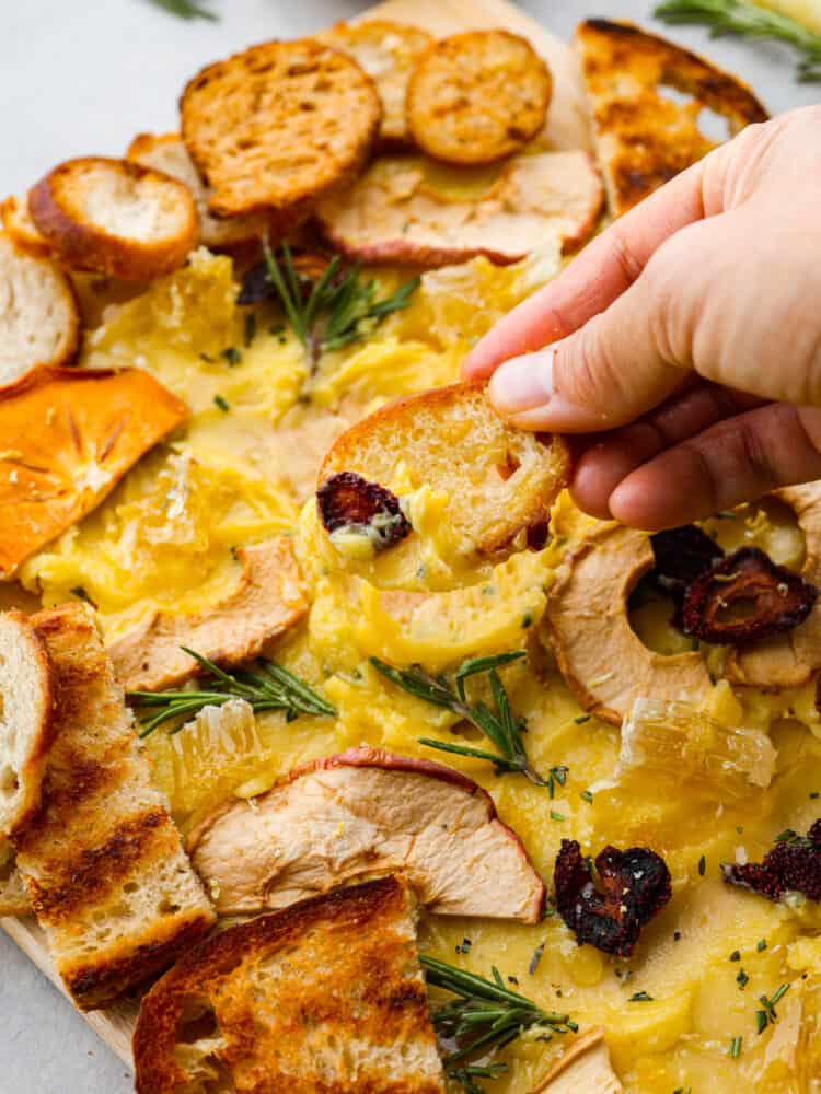 A butter board is the latest trend that you absolutely MUST TRY for your next gathering! This board is packed with the most delicious butter that is drizzled with honey and lemon zest and adds the perfect touch to your favorite bread! A hand scooping some butter onto a slice of crostini bread.