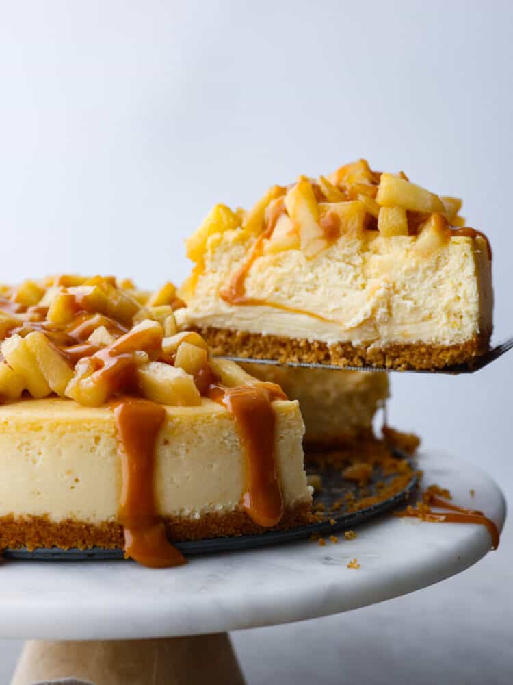 This Caramel Apple Cheesecake is the best of both desserts! Creamy tangy cheesecake topped with brown sugar apples and covered in caramel sauce, it's so crave-able you'll have a hard time eating just one slice. A side view of Caramel Apple Cheesecake. A slice being lifted out by a spatula.