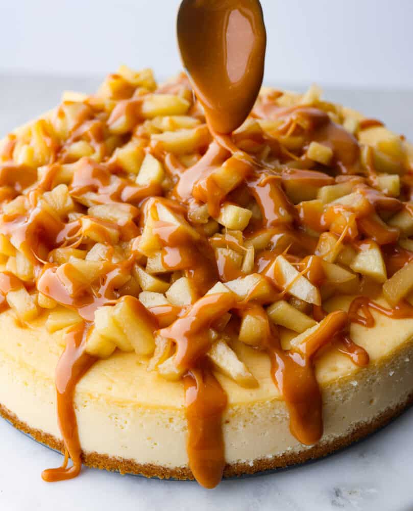 This Caramel Apple Cheesecake is the best of both desserts! Creamy tangy cheesecake topped with brown sugar apples and covered in caramel sauce, it's so crave-able you'll have a hard time eating just one slice. Caramel apple cheesecake with a spoon with caramel on it drizzling caramel over the top.