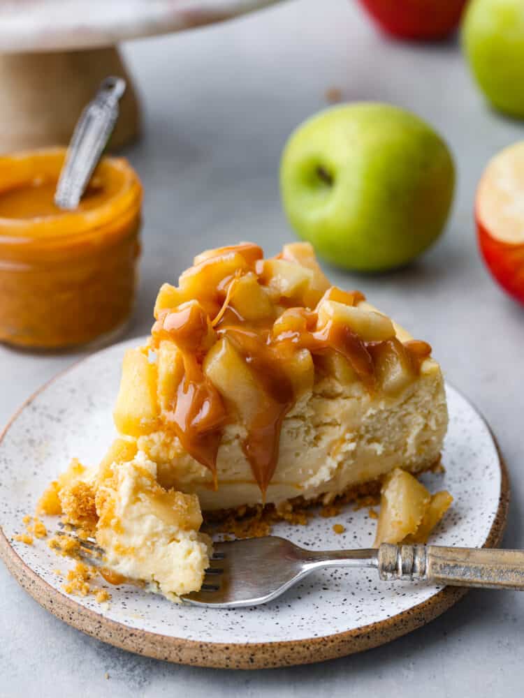 This Caramel Apple Cheesecake is the best of both desserts! Creamy tangy cheesecake topped with brown sugar apples and covered in caramel sauce, it's so crave-able you'll have a hard time eating just one slice. A slice of Caramel Apple Cheesecake with a bite taken out by a fork sitting on a stone ware plate with caramel and apples in the background.