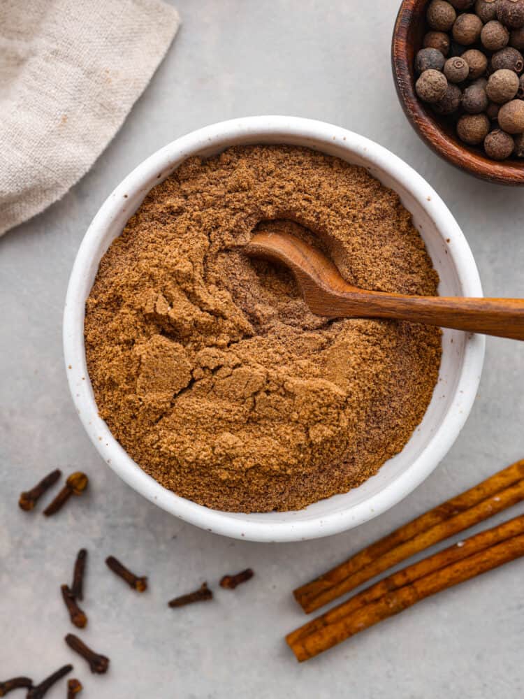There are so many great reasons to make Chai Spice at home! It is a blend of warm fall spices that make it perfect for cakes, cookies, and even your coffee or tea! Chai spice in a white bowl with a wooden spoon sitting in it with spices around it.