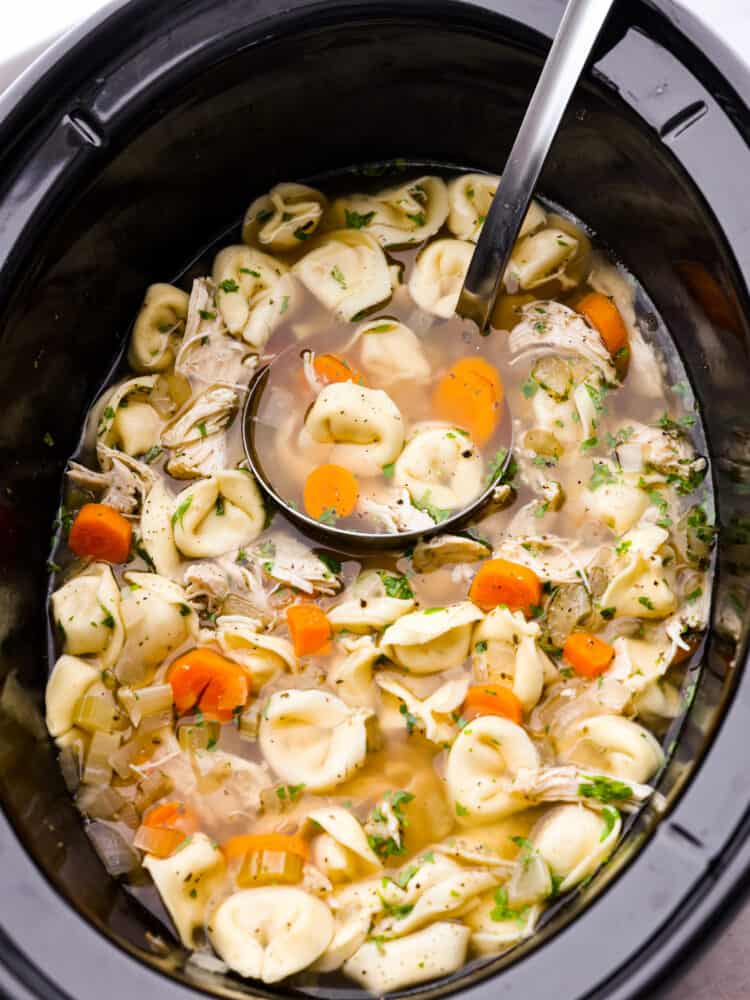 Slow cooker chicken tortellini soup is loaded with chopped vegetables, shredded cooked chicken, and cheesy tortellini all slow-cooked in a flavorfulย chicken broth. It's definitely a dinner the whole family will love! The top view of soup in a slow cooker with a label scooping some out.