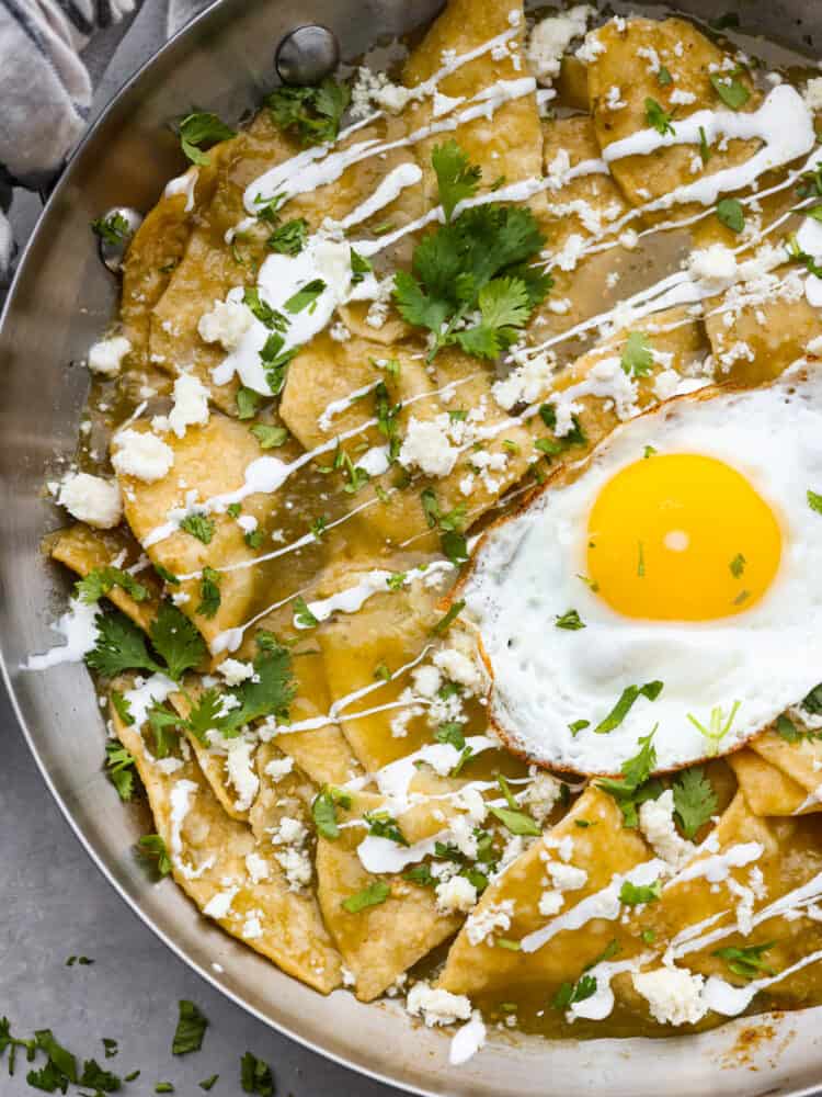 Fried tortilla chips drowning in salsa verde with your choice of toppings make this Chilaquiles recipe irresistible! Serve it with a fried egg and crema on top for a filling breakfast that is ready in minutes. You'll come back to this recipe again and again! A close up of Chilaquiles in a pan that shows the cilantro, crema, and cheese on top.