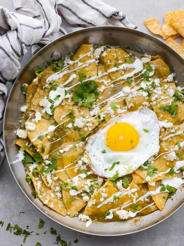 Fried tortilla chips drowning in salsa verde with your choice of toppings make this Chilaquiles recipe irresistible! Serve it with a fried egg and crema on top for a filling breakfast that is ready in minutes. You'll come back to this recipe again and again! Chilaquiles in a pan with a fried egg on top with a tea towel and tortilla chips in the background.
