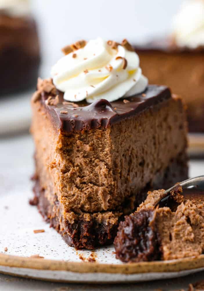 Chocolate brownie cheesecake - the ultimate cheesecake for chocolate lovers! It has a fudgy brownie bottom topped with a rich and creamy chocolate cheesecake, chocolate ganache, and whipped cream. Front view of a slice of chocolate cheesecake with a bite taken out of it.