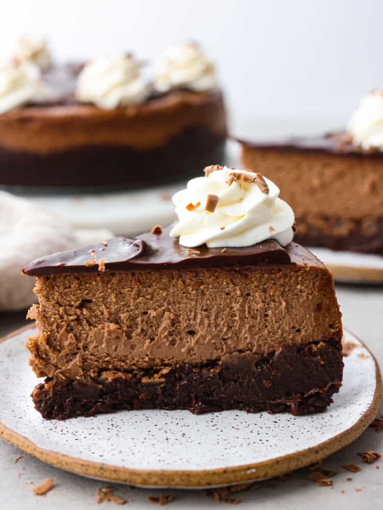 Chocolate brownie cheesecake - the ultimate cheesecake for chocolate lovers! It has a fudgy brownie bottom topped with a rich and creamy chocolate cheesecake, chocolate ganache, and whipped cream. A slice of brownie chocolate cheesecake on a stoneware plate.
