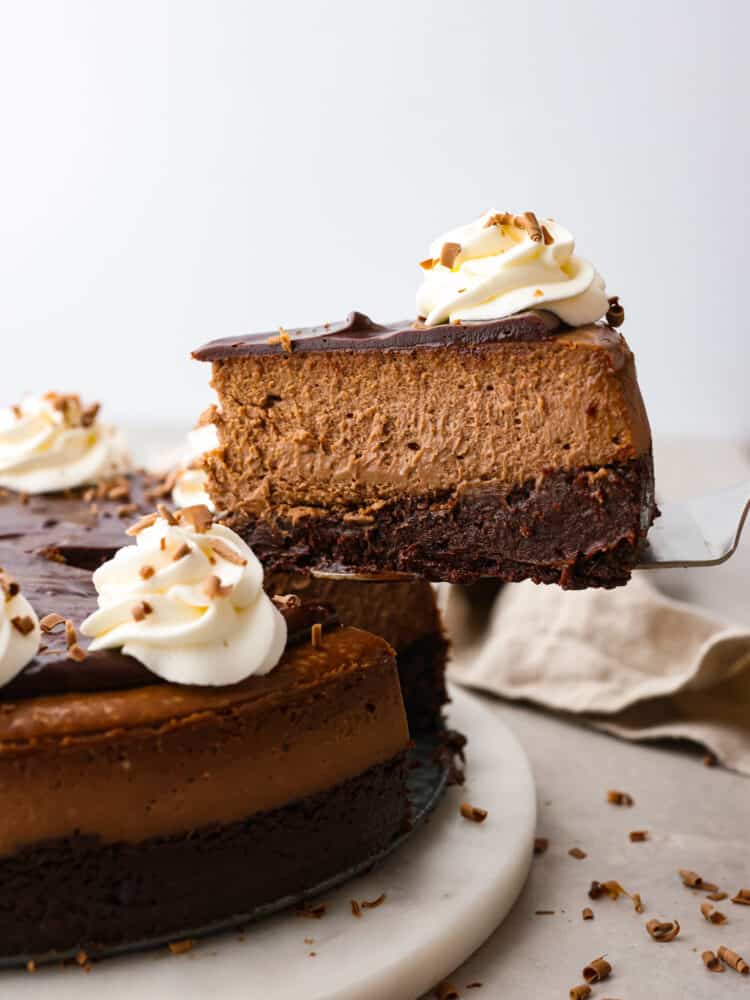 Chocolate brownie cheesecake - the ultimate cheesecake for chocolate lovers! It has a fudgy brownie bottom topped with a rich and creamy chocolate cheesecake, chocolate ganache, and whipped cream. A slice of cheesecake being served with a cake spatula.