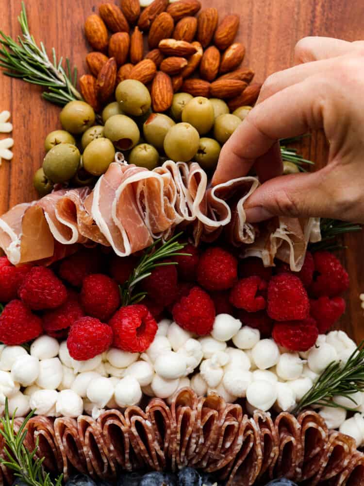 The cutest addition to any holiday party, this Christmas tree charcuterie board is both festive and delicious! It features a delicious mix of salami, prosciutto, cheese, fruits, and nuts, with rosemary sprigs as tree branches! It almost looks too good to eat. Adding prosciutto to the Christmas tree charcuterie.