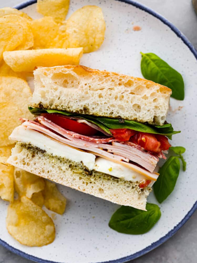 This Ciabatta Sandwich is a flavor packed and quick meal that is sure to satisfy! Fluffy and soft ciabatta bread is layered with quality deli turkey and salami on top of a bed of mozzarella and pesto for the perfect bite every time. Sandwich on a plate with potato chips and basil next to it.