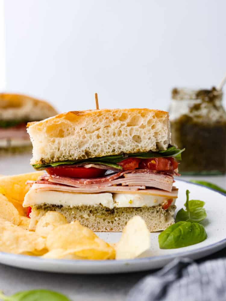 This Ciabatta Sandwich is a flavor packed and quick meal that is sure to satisfy! Fluffy and soft ciabatta bread is layered with quality deli turkey and salami on top of a bed of mozzarella and pesto for the perfect bite every time. Ciabatta sandwich on a white plate with potato chips and basil next to it.