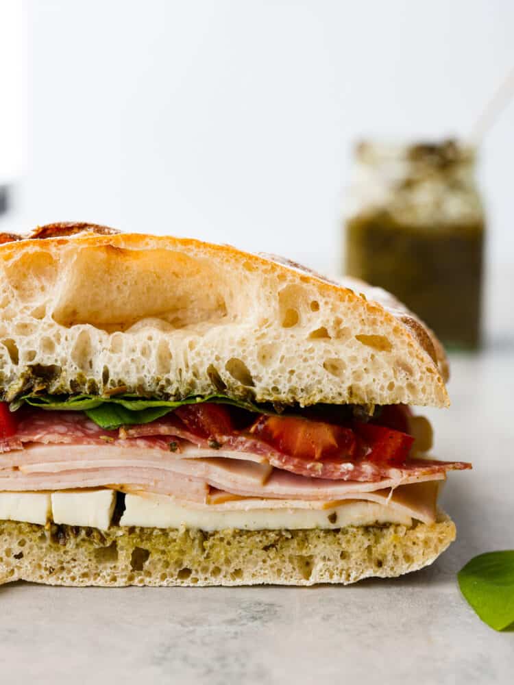 This Ciabatta Sandwich is a flavor packed and quick meal that is sure to satisfy! Fluffy and soft ciabatta bread is layered with quality deli turkey and salami on top of a bed of mozzarella and pesto for the perfect bite every time. Ciabatta sandwich on a counter top with pesto in the background.
