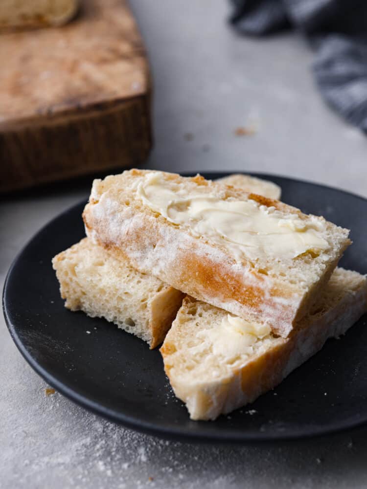 Make this classic Italian ciabatta bread recipe at home with just 4 simple ingredients! Crusty on the outside, and perfectly chewy on the inside, this bread is delicious for sandwiches, served with soup, or simply toasted. Close up view of sliced and buttered bread on a black plate. A wood cutting board nearby with scattered crumbs around the plate.