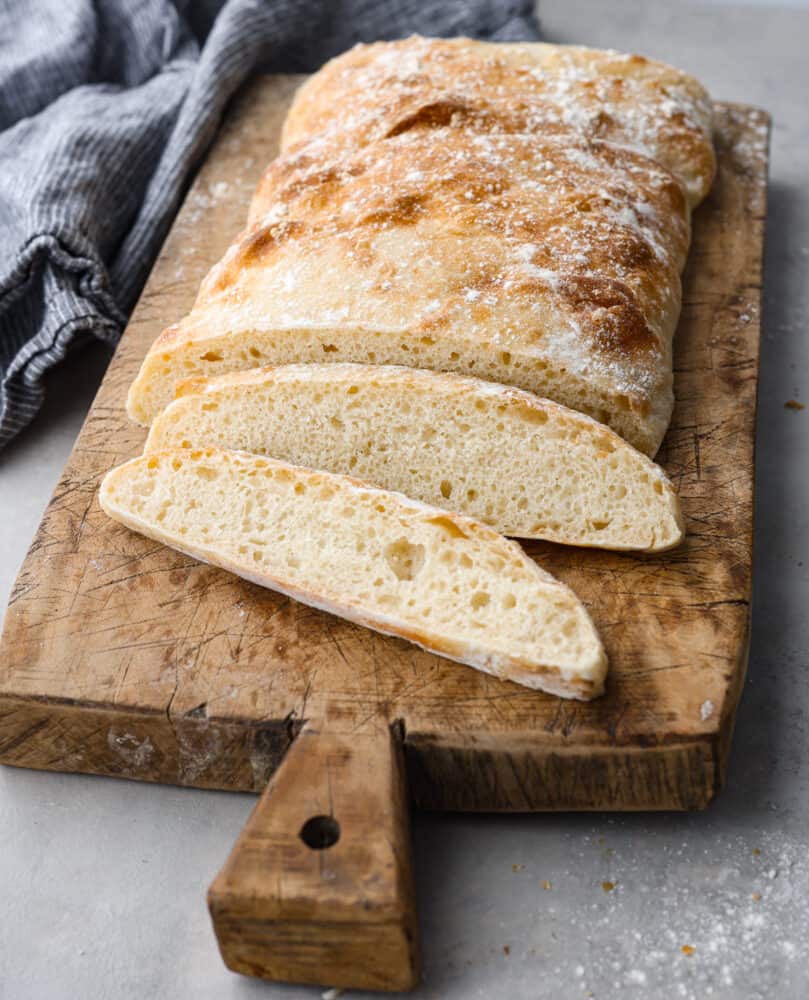 Make this classic Italian ciabatta bread recipe at home with just 4 simple ingredients! Crusty on the outside, and perfectly chewy on the inside, this bread is delicious for sandwiches, served with soup, or simply toasted. Side view of baked and slice ciabatta bread on a wood cutting board. A blue kitchen towel is next to the wood board.