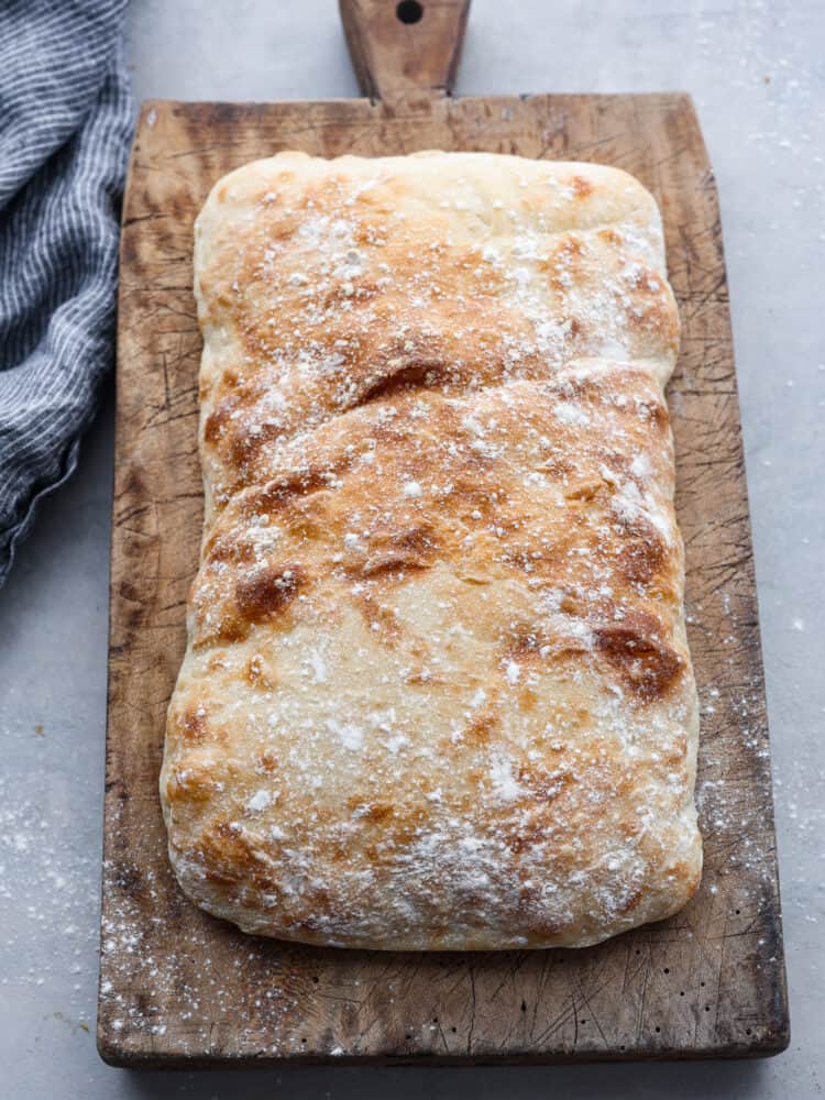 Make this classic Italian ciabatta bread recipe at home with just 4 simple ingredients! Crusty on the outside, and perfectly chewy on the inside, this bread is delicious for sandwiches, served with soup, or simply toasted. Top view of a loaf of ciabatta bread on a wood cutting board. A blue kitchen towel is next to the wood board. Flour is sprinkled on top of the loaf.