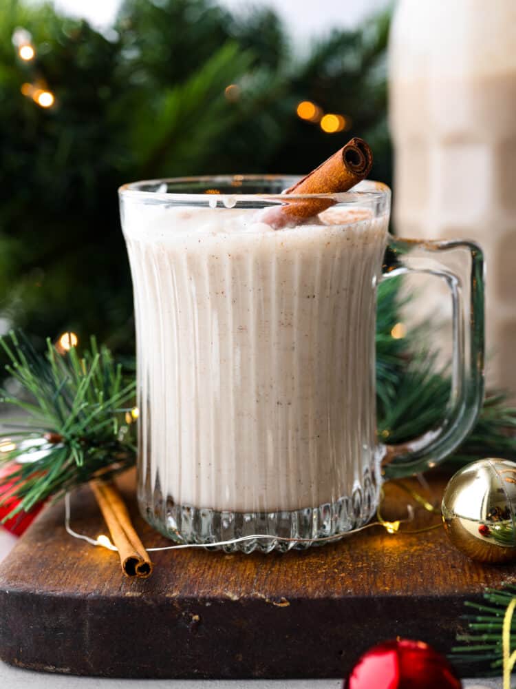 Coquito is a seasonal Puerto Rican drink that has coconut milk and cream with warm spices like cinnamon and nutmeg. Welcome in the holiday season with this unique and festive drink. It will become a new holiday tradition! Coquito with a cinnamon stick in it and green branches behind it.