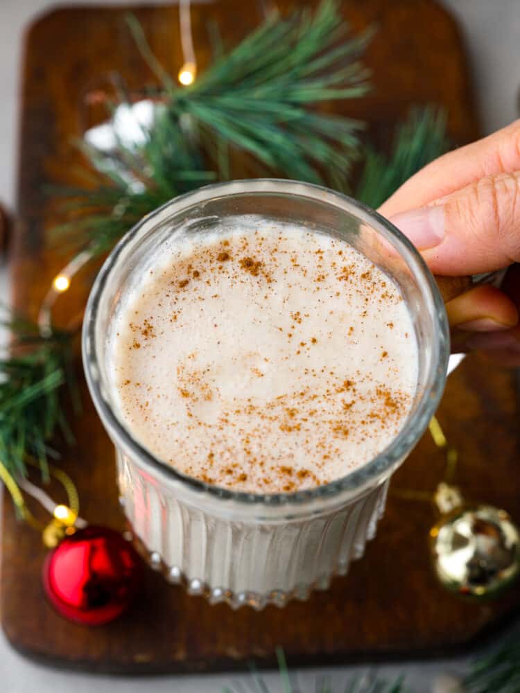 Coquito is a seasonal Puerto Rican drink that has coconut milk and cream with warm spices like cinnamon and nutmeg. Welcome in the holiday season with this unique and festive drink. It will become a new holiday tradition! A hand holding coquito over a table.