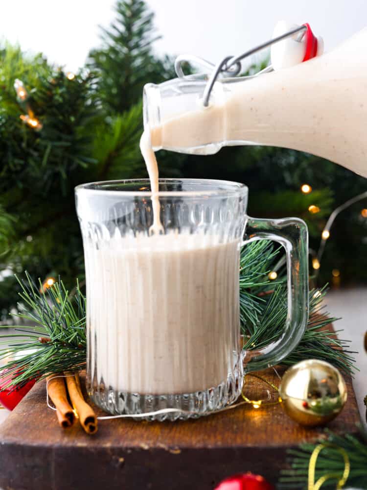 Coquito is a seasonal Puerto Rican drink that has coconut milk and cream with warm spices like cinnamon and nutmeg. Welcome in the holiday season with this unique and festive drink. It will become a new holiday tradition! Coquito being poured into a glass with green branches and lights behind it.